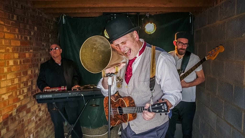 A man with a ukelele sings into a brass megaphone - a keyboard player and bass player stand in the background