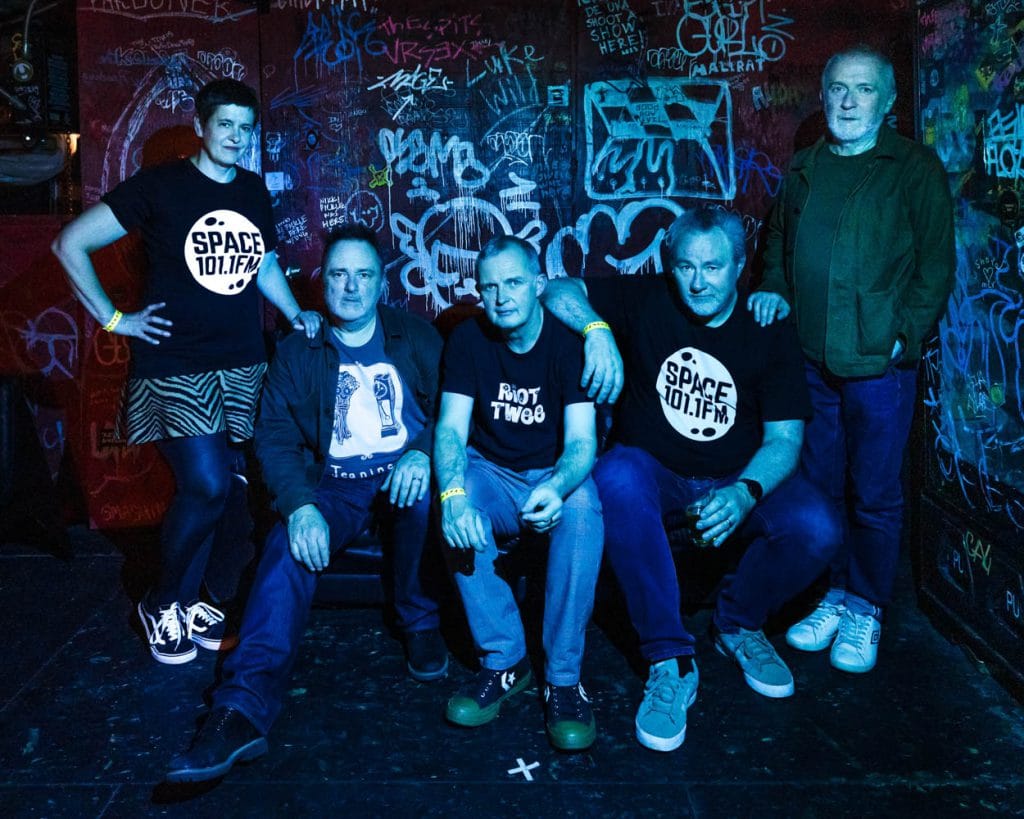 Five musicians lined up against a wall covered in graffiti